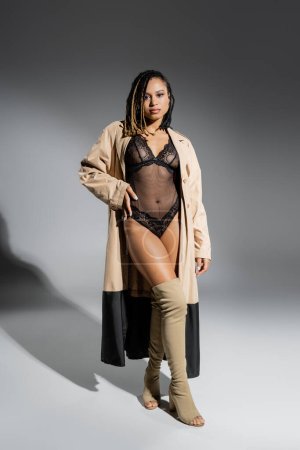 longitud completa de provocativa y atractiva mujer afroamericana en traje de encaje negro, gabardina beige y botas de rodilla posando con la mano en la cadera y mirando a la cámara sobre fondo gris