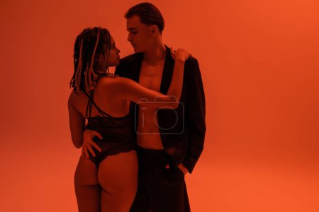junger und stilvoller Mann, der mit der Hand in der Tasche schwarzer Shorts steht und eine attraktive Afroamerikanerin mit sexy Gesäß umarmt, trägt einen Spitzenbody auf orangefarbenem Hintergrund mit roter Beleuchtung