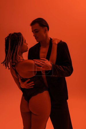 young and stylish man in black blazer hugging appealing african american woman with dreadlocks and sexy buttocks, wearing lace bodysuit on orange background with red lighting effect