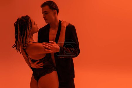 Photo for Young and handsome man in stylish blazer embracing alluring african american man with sexy buttocks wearing lace bodysuit on orange background with red lighting effect - Royalty Free Image