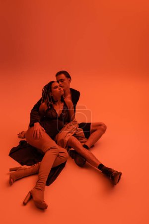 full length of passionate interracial couple, african american woman in black lace bodysuit with over knee boots, and young man sitting on clothes on orange background with red lighting effect