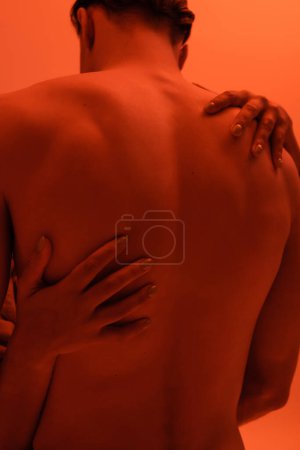 Rückansicht des jungen, hemdlosen und sexy Mannes in der Nähe einer leidenschaftlichen Afroamerikanerin, die seinen muskulösen Körper auf orangefarbenem Hintergrund mit rotem Lichteffekt umarmt