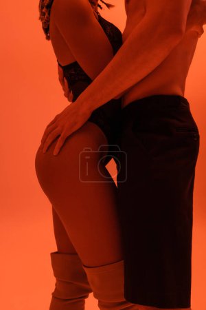 Foto de Vista parcial de sexy pareja interracial, hombre sin camisa en pantalones negros y mujer afroamericana en traje de encaje abrazando sobre fondo naranja con efecto de iluminación roja - Imagen libre de derechos