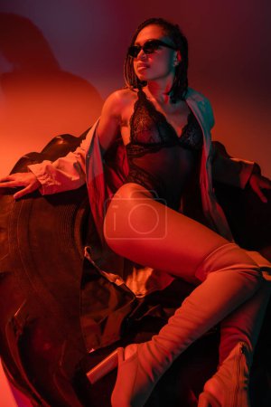 Photo for Fashionable and sexy african american woman in sunglasses, black lace bodysuit, beige trench coat and over knee boots sitting on huge tire on grey background with orange lighting - Royalty Free Image