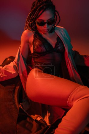 freche und sexy afrikanisch-amerikanische Frau mit dunkler stylischer Sonnenbrille, schwarzem Spitzenbody und beigem Trenchcoat sitzt auf einem riesigen Reifen auf grauem Hintergrund mit roter Beleuchtung