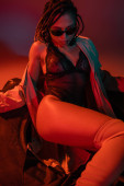 sassy and sexy african american woman in dark stylish sunglasses, black lace bodysuit and beige trench coat sitting on huge tire on grey background with red lighting mug #658774952
