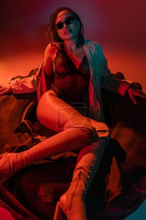 Photo for Provocative african american woman in dark trendy sunglasses, black lace bodysuit, beige trench coat and over knee boots posing on huge tire on grey background with red lighting - Royalty Free Image