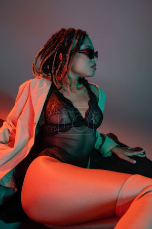 seductive african american woman with dreadlocks wearing dark sunglasses, black lace bodysuit and beige trench coat sitting on huge tire and looking away on grey background with red lighting