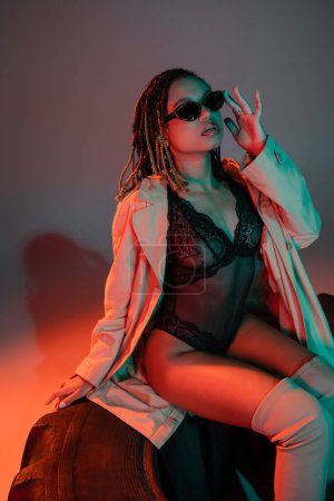 mujer afroamericana glamorosa y atrevida ajustando gafas de sol oscuras y elegantes mientras está sentada sobre un enorme neumático en traje de encaje negro y abrigo beige sobre fondo gris con iluminación roja
