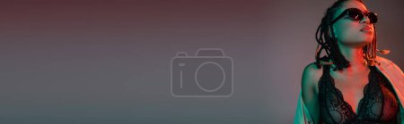 Téléchargez les photos : Femme afro-américaine passionnée et élégante avec dreadlocks posant dans des lunettes de soleil sombres, lingerie en dentelle noire et trench coat beige sur fond gris avec éclairage rouge, bannière - en image libre de droit