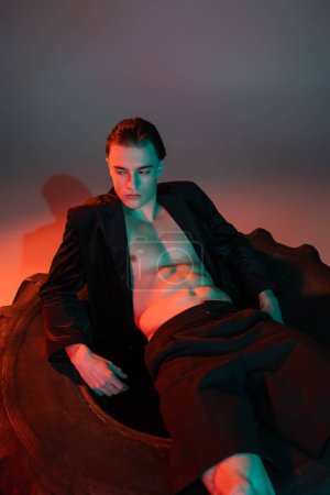 sexy and fashionable man with brunette hair, wearing black blazer on shirtless muscular body, sitting on huge tire and looking away on grey background with red lighting
