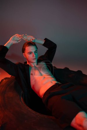 confident and sexy man in black trendy blazer on shirtless muscular torso posing on huge tire with hands above head on grey background with red lighting