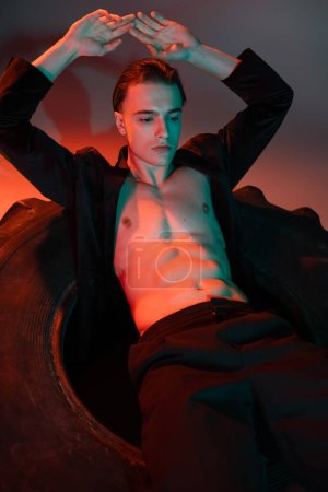 Téléchargez les photos : Jeune et sexy homme portant blazer noir sur torse musclé torse nu, assis sur un énorme pneu et posant avec les mains au-dessus de la tête sur fond gris avec éclairage rouge - en image libre de droit