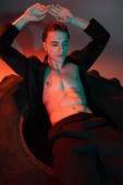 young and sexy man wearing black blazer on shirtless muscular torso, sitting on huge tire and posing with hands above head on grey background with red lighting puzzle #658775218