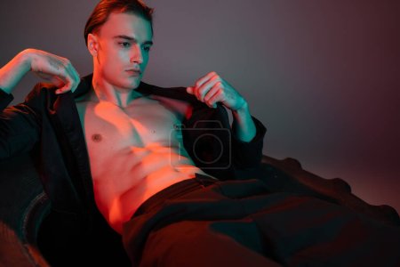 handsome and seductive man with brunette hair posing on huge tire in black stylish blazer on shirtless muscular torso on grey background with red lighting