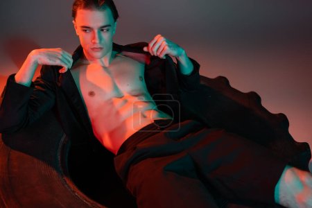 young, self-assured and charismatic man in black fashionable blazer on shirtless muscular body sitting on huge tire and looking away on grey background with red lighting