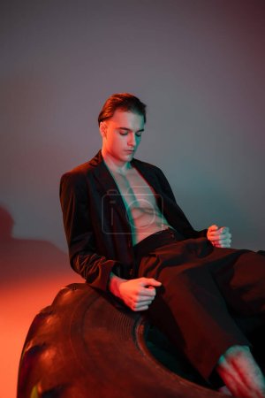 junger, gut aussehender und sexy Mann im schwarzen trendigen Blazer auf hemdlosem muskulösem Oberkörper sitzt und posiert auf riesigen Reifen auf grauem Hintergrund mit roter Beleuchtung