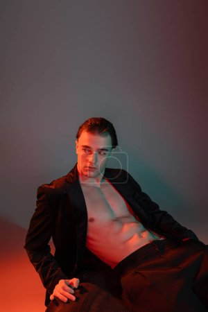 young, handsome and sexy man in black stylish blazer on shirtless muscular body sitting on huge tire and looking away on grey background with red lighting