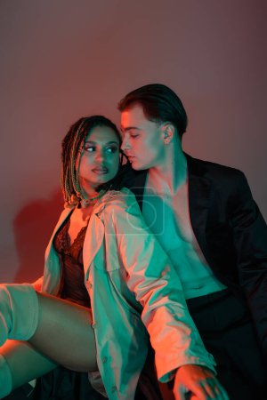 Photo for Confident and stylish man in black blazer sitting near african american woman with dreadlocks wearing lace bodysuit and beige trench coat on grey background with red lighting - Royalty Free Image