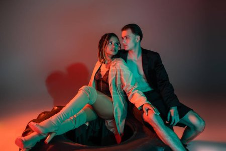 Sexy african american woman in black lace bodysuit, beige trench coat and over knee boots sitting on huge tire and looking at charismatic man in stylish blazer on grey background with red lighting