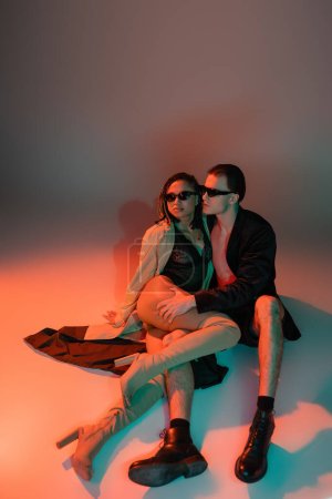 full length of glamour man in dark sunglasses and black blazer hugging leg of sassy african american woman in lace bodysuit, beige trench coat and over knee boots on grey with red lighting