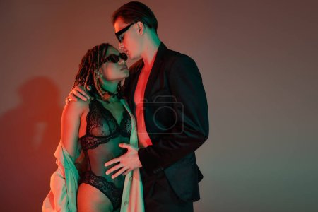 glamorous man in dark sunglasses and blazer embracing provocative african american woman in black lace bodysuit and beige trench coat on grey background with red lighting