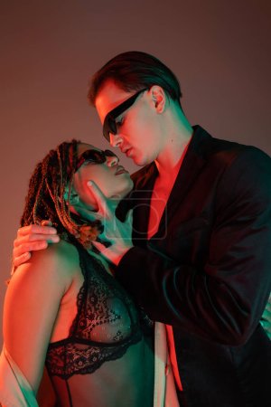 young and self-assured man in dark sunglasses and black blazer seducing enchanting african american woman in lace bodysuit on grey background with red lighting