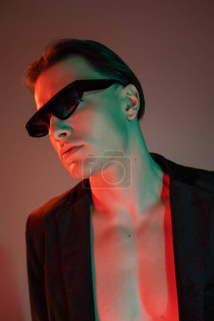 portrait of young bare-chested man in black fashionable blazer and dark stylish sunglasses looking away while posing on grey background with red lighting
