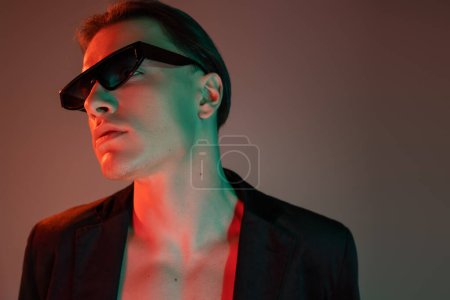 young, self-assured and bare-chested man in black blazer and dark fashionable sunglasses standing and looking away on grey background with red lighting