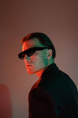 ausdrucksstarker und selbstbewusster Mann mit brünetten Haaren, dunkler trendiger Sonnenbrille und schwarzem Blazer, der im Stehen wegschaut und auf grauem Hintergrund mit roter Beleuchtung posiert