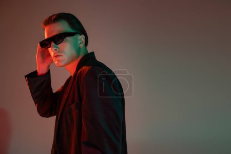 Téléchargez les photos : Jeune homme à la mode et confiant ajustant des lunettes de soleil sombres et élégantes tout en se tenant debout et posant en blazer noir sur fond gris avec éclairage rouge et espace de copie - en image libre de droit