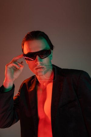 young, bare-chested and appealing man in black blazer adjusting dark fashionable sunglasses and looking at camera on grey background with red lighting