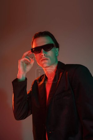joven y guapo hombre con cabello moreno ajustando gafas de sol oscuras de moda mientras posan en chaqueta negra y mirando a la cámara sobre fondo gris con iluminación roja