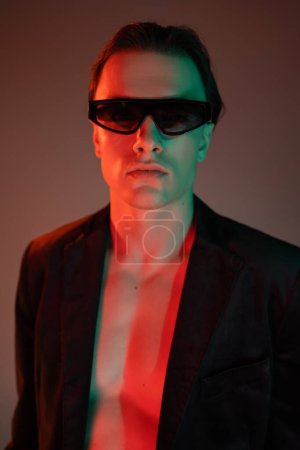 portrait of young and fashionable man in dark sunglasses and stylish blazer looking at camera while standing and posing on grey background with red lighting