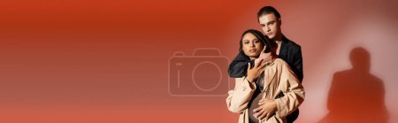 Photo for Young and trendy man in blazer hugging charming african american woman in black lace bodysuit and beige trench coat while looking at camera on red and pink background, banner - Royalty Free Image