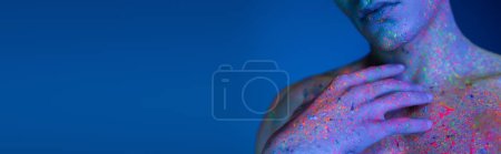 Photo for Partial view of youthful man holding hand near bare chest while posing in radiant and multicolored neon body paint on blue background with cyan lighting effect, banner - Royalty Free Image