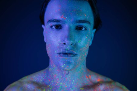 Photo for Portrait of confident, self-assured man with bare shoulders posing in vibrant and colorful neon body paint while looking at camera on blue background with cyan lighting effect - Royalty Free Image