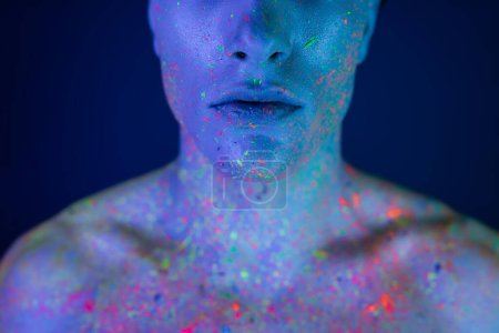 Teilaufnahme eines jungen, hemdlosen Mannes in buntem und lebendigem Neon-Body-Paint stehend und posierend auf verschwommenem blauem Hintergrund mit Cyan-Lichteffekt