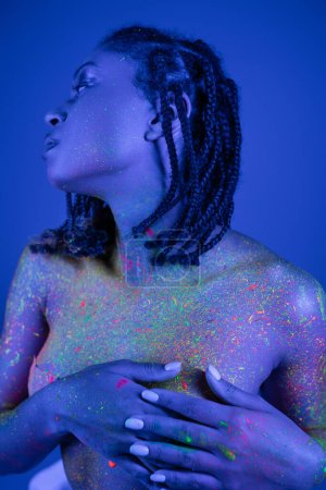 young, sensual and bare-chested african american woman with dreadlocks covering breast with hands while posing in colorful neon body paint on blue background with cyan lighting effect