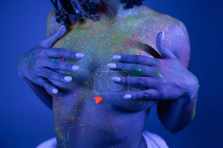 Teilaufnahme einer jugendlichen und barbusigen afrikanisch-amerikanischen Frau in lebendiger und farbenfroher Neon-Körperfarbe, die die Brust mit Händen auf blauem Hintergrund mit Cyan-Lichteffekt bedeckt