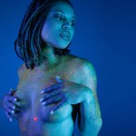 charming and nude african american woman posing in radiant and colorful neon body paint while looking away and covering breast with hands on blue background with cyan lighting effect