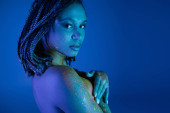 intriguing african american woman in multicolored neon body paint looking at camera and covering breast with hands while posing on blue background with cyan lighting effect puzzle #658776154