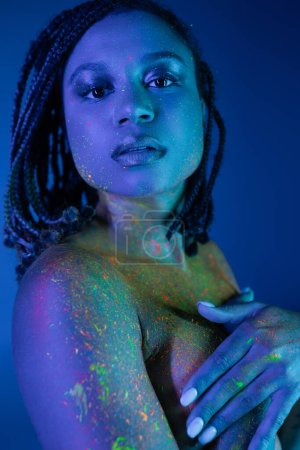 expressive and charming african american woman in radiant and multicolored neon body paint covering breast with hands and looking at camera on blue background with cyan lighting effect