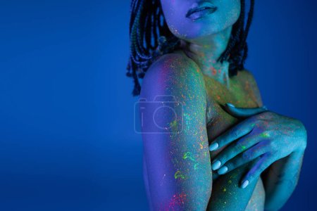 Partial view of nude african american woman in bright and colorful neon body paint covering breast with hands while standing on blue background with cyan lighting effect
