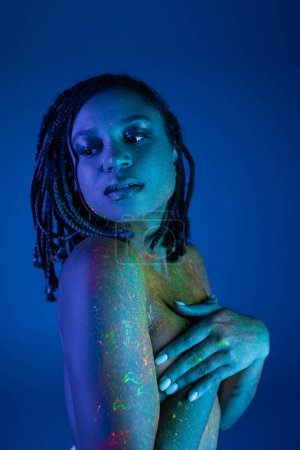 young and tempting bare-chested african american woman with dreadlocks, in colorful neon body paint, covering breast with hands and looking away on blue background with cyan lighting effect