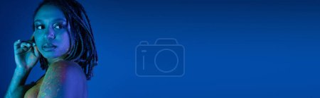 Téléchargez les photos : Femme américaine africaine sensuelle avec dreadlocks et épaule nue, dans la peinture au néon colorée rayonnante du corps regardant loin sur fond bleu avec effet d'éclairage cyan, bannière - en image libre de droit