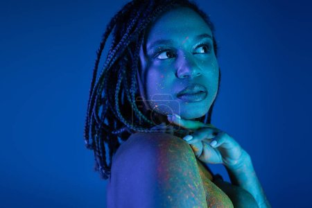 Porträt einer bezaubernden und sexy afrikanisch-amerikanischen Frau mit Dreadlocks, in farbenfroher Neon-Körperfarbe, die wegschaut, während sie die Hand am Kinn hält, auf blauem Hintergrund mit Cyan-Lichteffekt