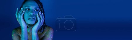 retrato de mujer afroamericana joven con rastas, en colorida pintura de cuerpo de neón, cogidas de la mano cerca de la cara y mirando hacia otro lado sobre fondo azul con efecto de iluminación cian, pancarta