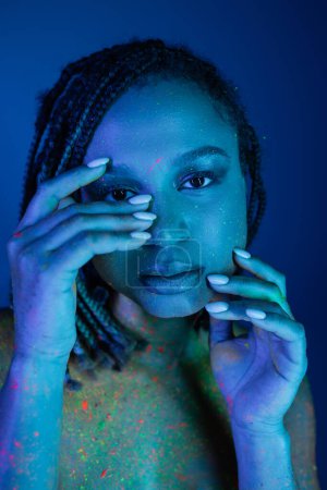 Porträt einer jungen und verführerischen afrikanisch-amerikanischen Frau mit Dreadlocks, die die Hände vor dem Gesicht hält, während sie in bunter Neon-Körperfarbe auf blauem Hintergrund mit Cyan-Lichteffekt posiert