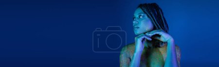 Photo for Sexy and eye-catching african american woman with dreadlocks, in multicolored neon body paint, holding hands near chin and looking away on blue background with cyan lighting effect, banner - Royalty Free Image